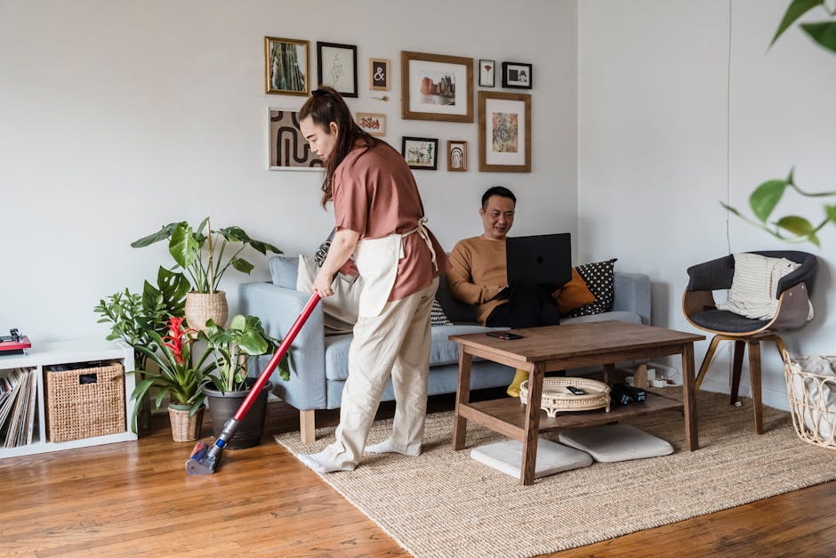 Woman vacuuming living room while man on laptop, modern home setting.